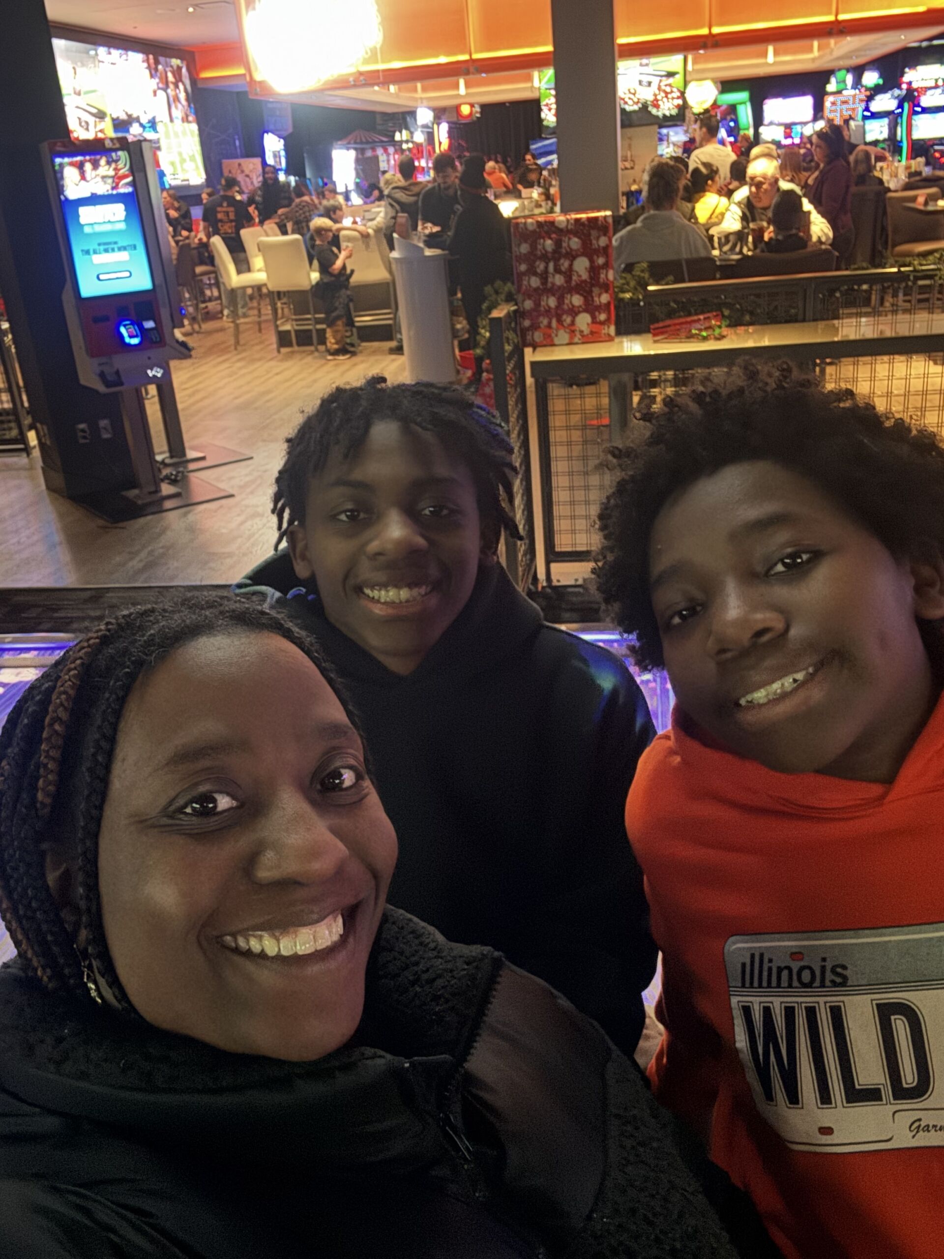 Erin KA with Lem & Aaron Kelley-Austin poses for a selfie with her two boys, Lem and Aaron, at a skating rink. They are smiling and you can see a crowd of other families behind them, sitting at tables. Her kids are teenage Black boys. Lem has short black locs and is wearing a black hoodie. Aaron has his hair out in a medium-length natural style and is wearing a bright orange hoodie that has words cut-off on it but you can clearly read "Illinois Wild" on it with a license plate-like design. Kelley-Austin has long black braids in her hair and is wearing a black winter coat.