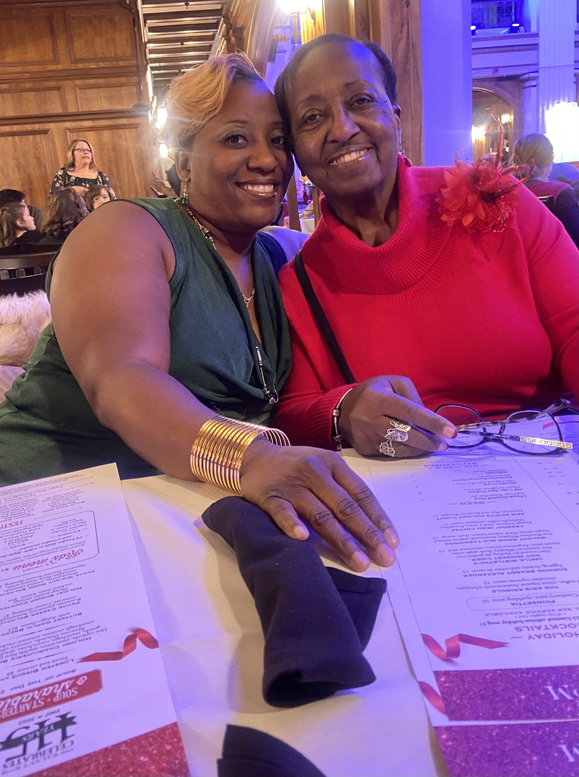 Erin KA with mother Kelley-Austin sits next to her mom at a fancy dinner table. They are dressed up and smiling brightly while leaning on each other, clearly celebrating. They are both Black woman: Kelley-Austin has short wavy blonde hair and is wearing a green blouse. Her mom has very short dark brown hair and is wearing a bright red blouse.