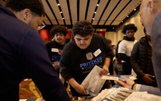A student in a Pritzker shirt leans over a table and in between two college representatives. He holds a packet in his hand as one of the reps hands him a pamphlet. You can see other students milling in the background.